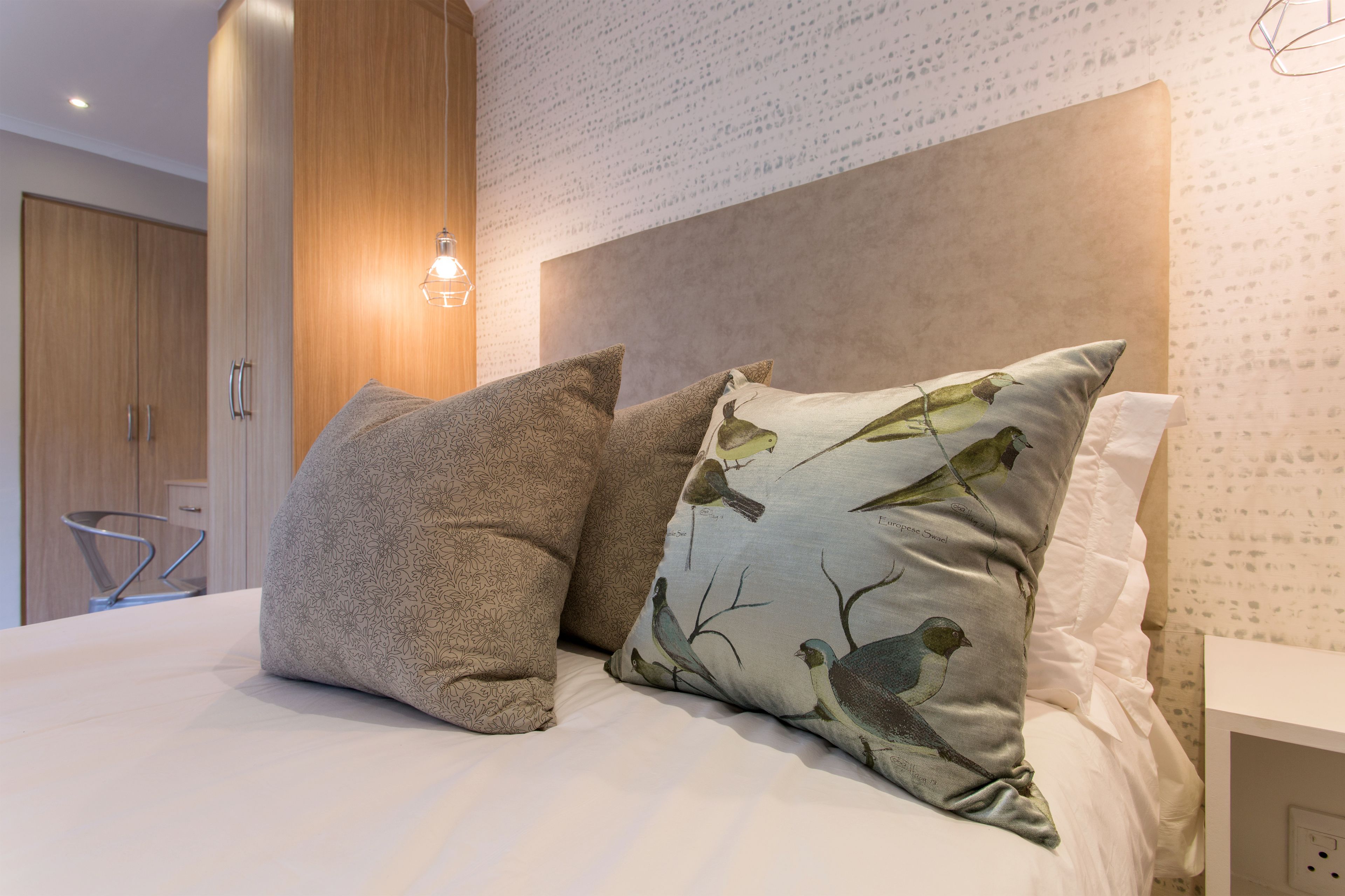 A neatly made bed with patterned pillows, a wooden wardrobe, and a wall with a subtle pattern.
