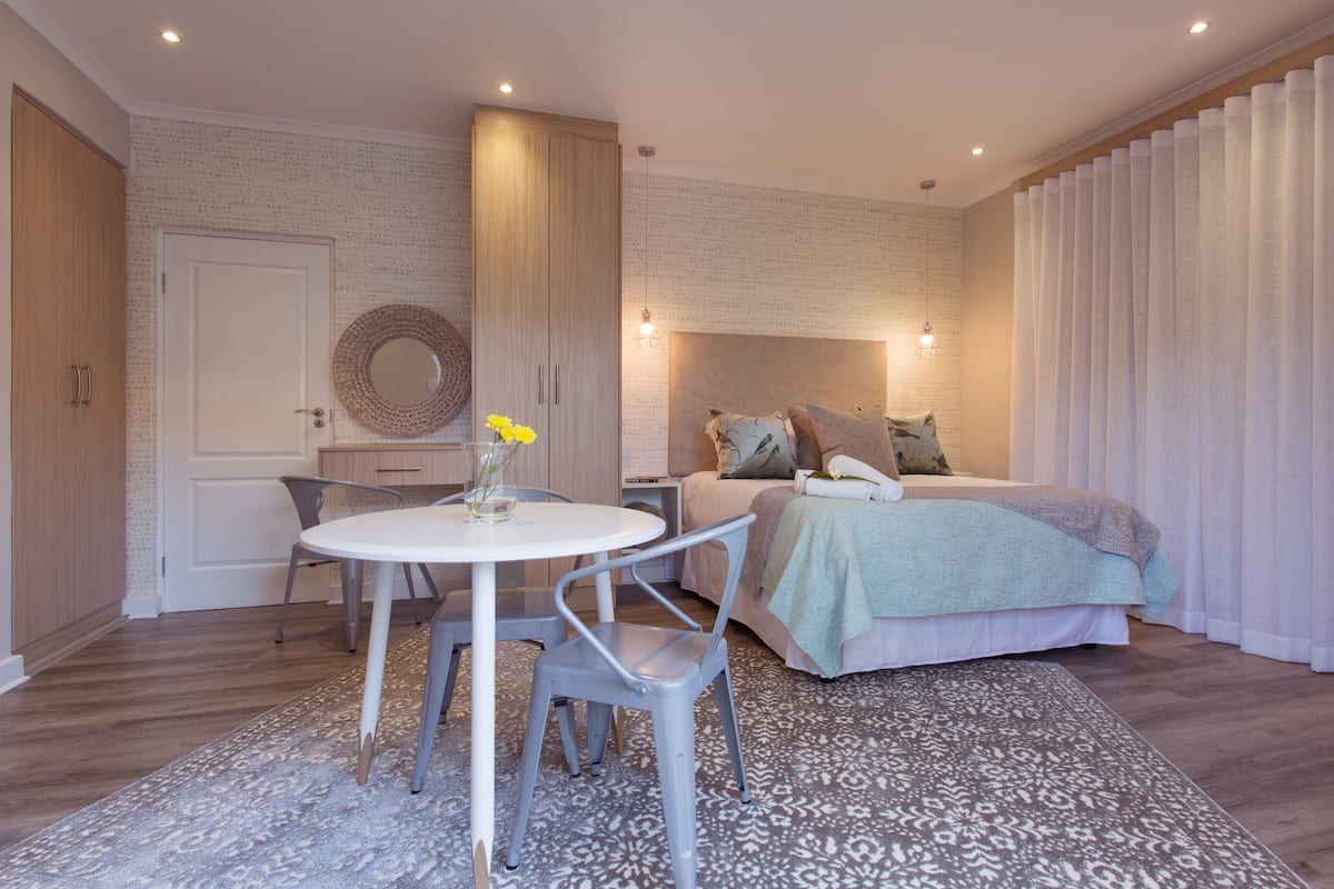 Un dormitorio moderno con cama, mesita de noche y un espejo redondo. Hay una mesa de comedor con sillas y una alfombra estampada.