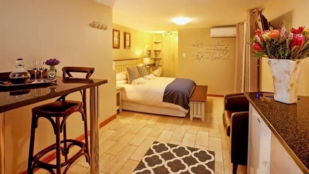 A compact room with a bed, a small table, and a chair. There is a wall with decorative text and a vase with flowers.