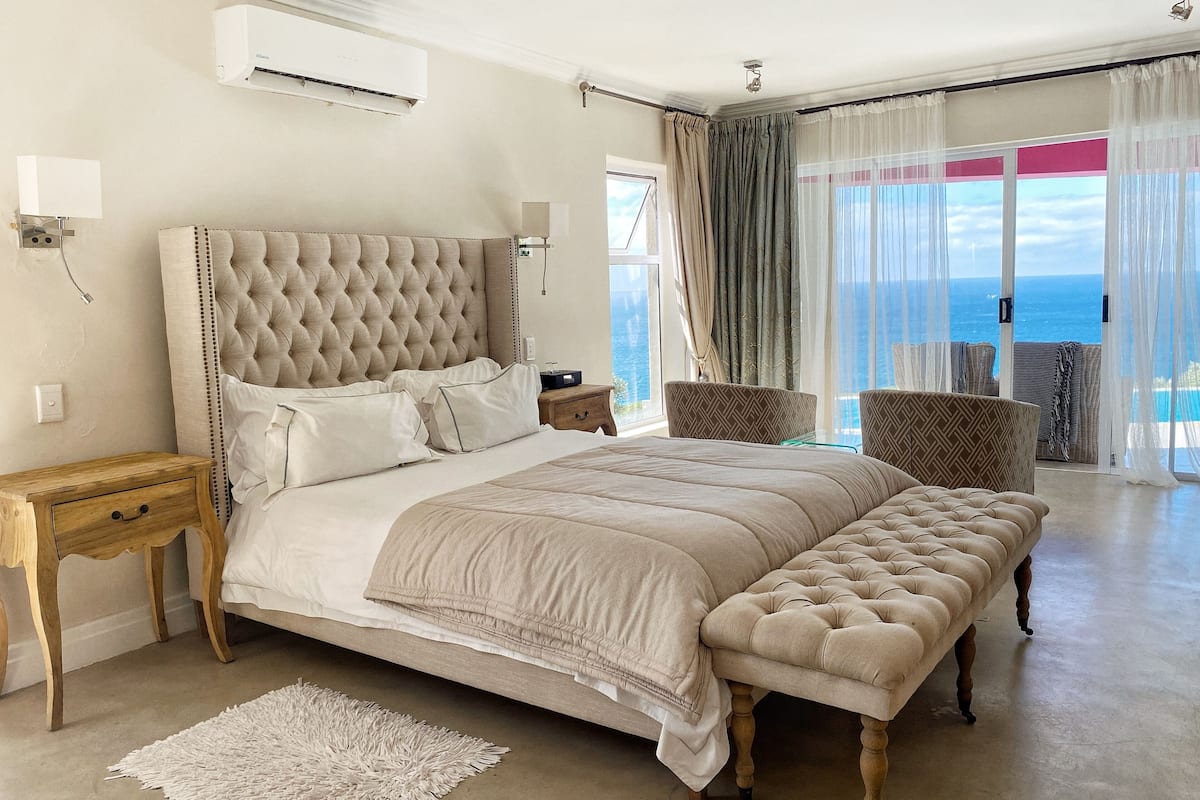 A spacious bedroom with a large bed, a wooden nightstand, a bench, and a view of the sea through large windows.