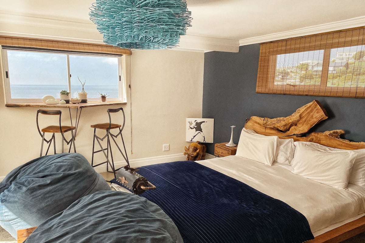A bedroom with a bed, a window with a view, two chairs, a wooden headboard, and a hanging light fixture.