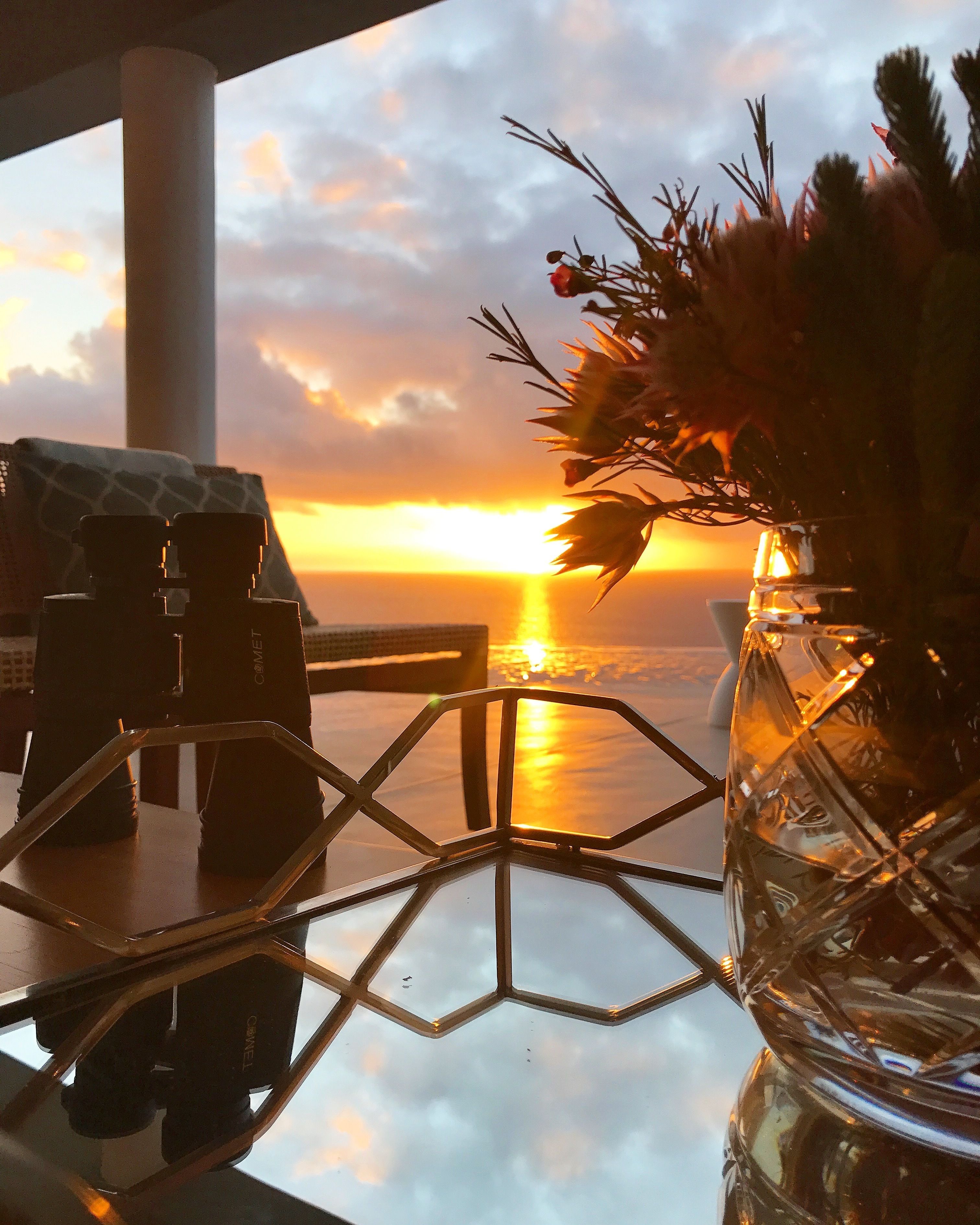 Una mesa con binoculares y un florero, con vista a una puesta de sol sobre el agua.