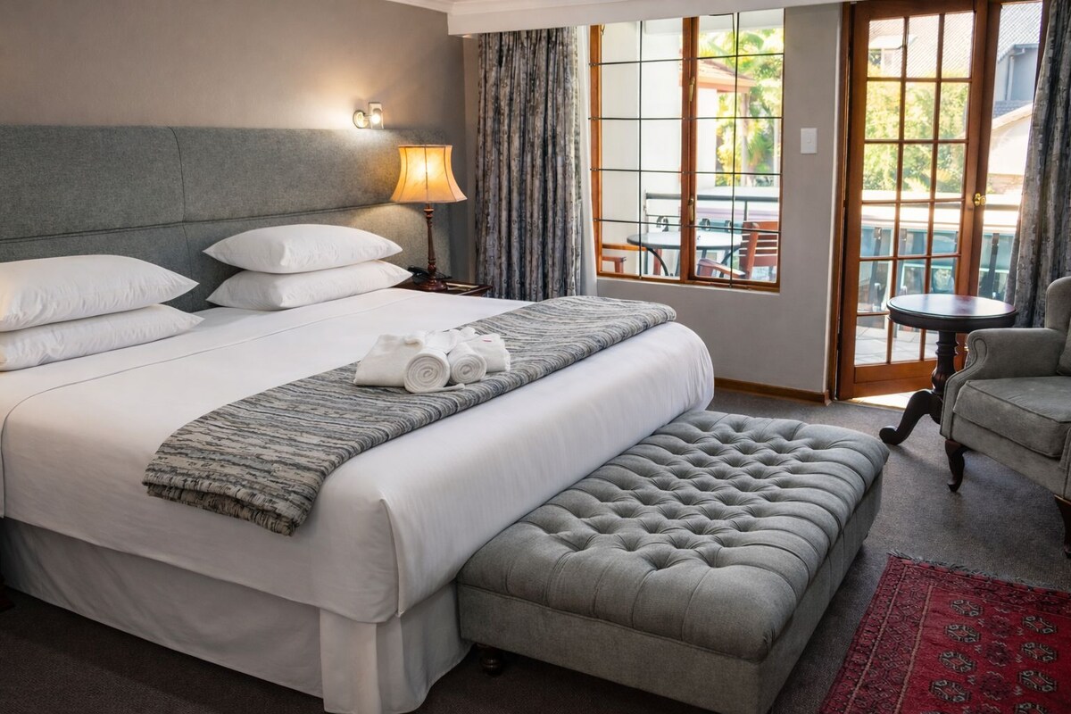 A hotel room with a large bed, a bedside lamp, a window with curtains, a small table, and a chair.