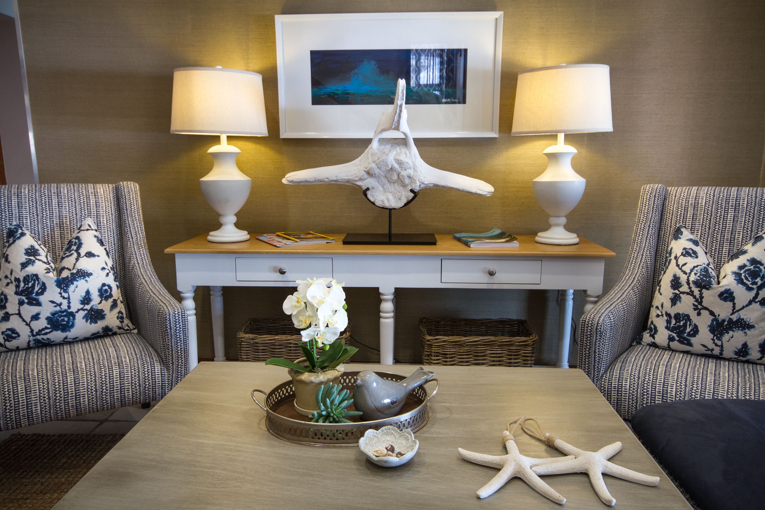 Una sala de estar con dos sillones, una mesa de centro con objetos decorativos, una obra de arte enmarcada en la pared y dos lámparas.