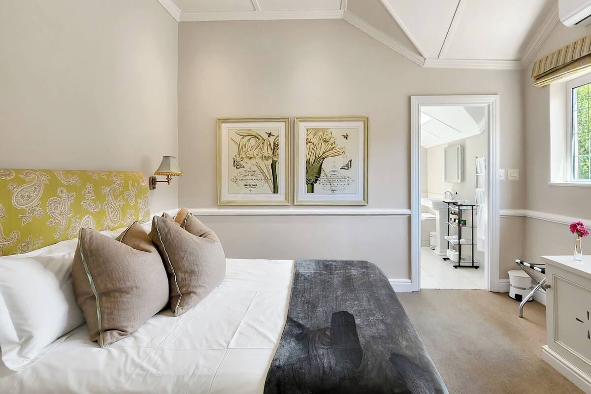 A bedroom with a bed, pillows, a bedside table, a mirror, and two framed botanical prints on the wall.