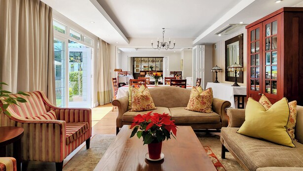 A spacious living room with a large sofa, a striped armchair, and a wooden coffee table. There is a dining area in the background with a chandelier. - Roodenburg House 1882 (Cape Town)
