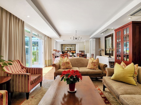 A spacious living room with a large sofa, a striped armchair, and a wooden coffee table. There is a dining area in the background with a chandelier.