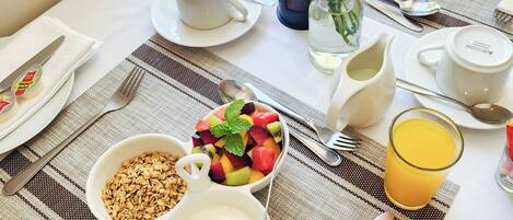 Una mesa de desayuno con un tazón de cereal, yogur, una ensalada de frutas, un vaso de jugo de naranja y un florero con flores.