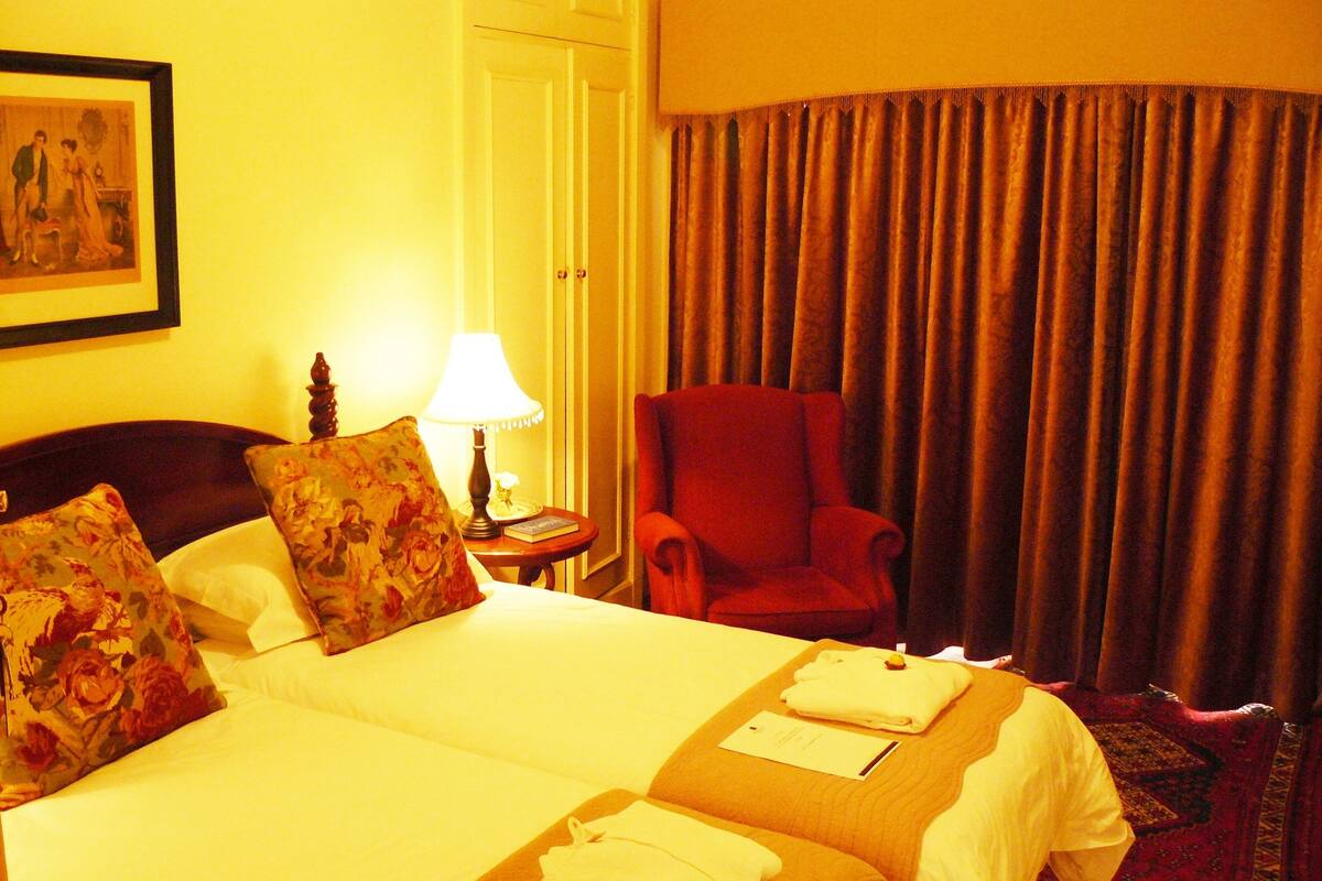 Habitación de hotel con una cama, dos almohadas, un sillón rojo, una lámpara y un cuadro en la pared.