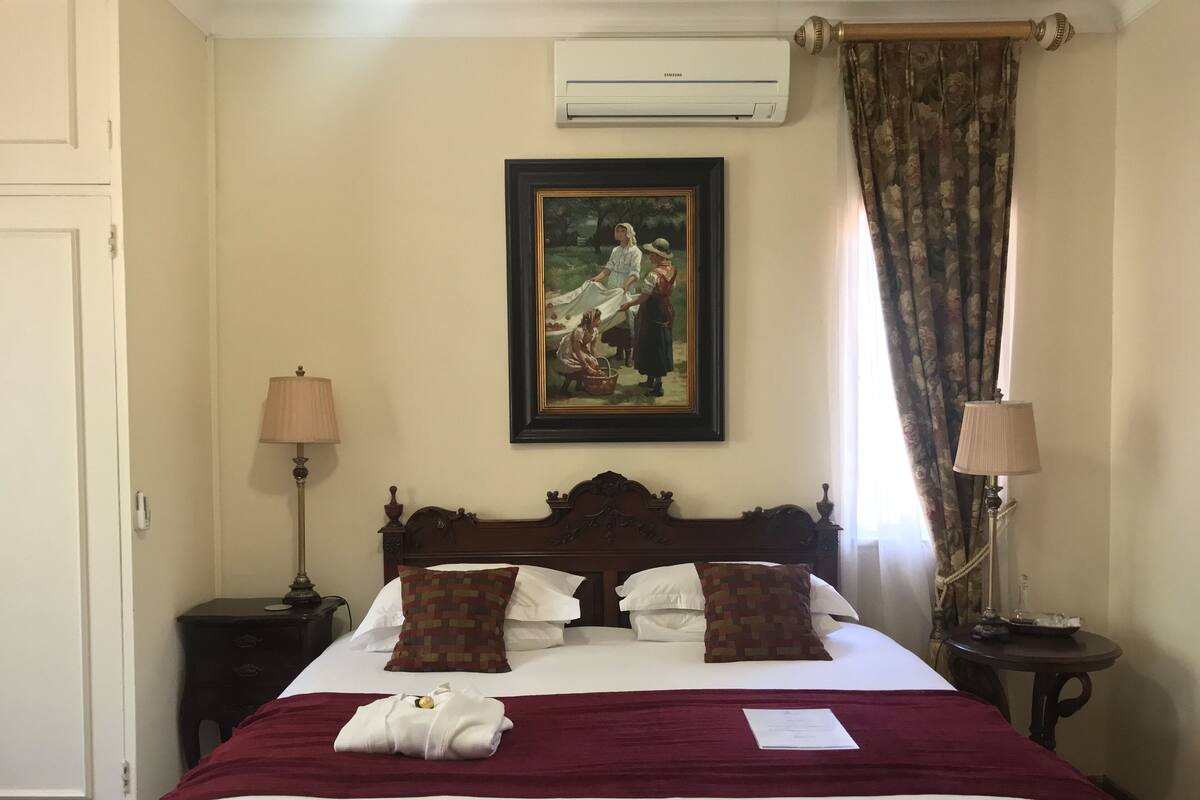 Una cama bien hecha con una manta roja, almohadas blancas y una toalla doblada. Encima de la cama hay un cuadro enmarcado, una lámpara a cada lado y un equipo de aire acondicionado en la pared.