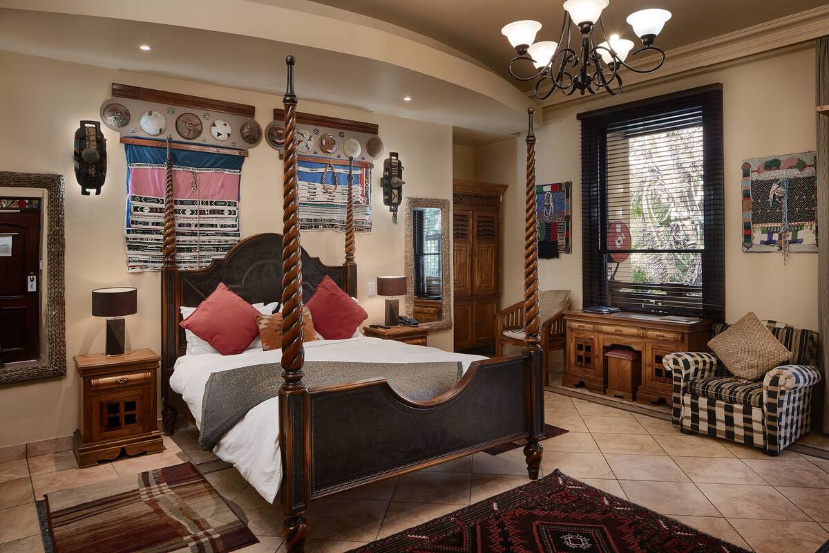 A four-poster bed with a wooden headboard, a wooden nightstand, a plaid-patterned chair, and a large window with blinds.