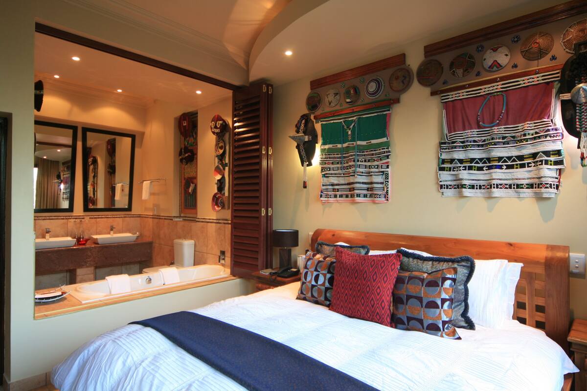 A hotel room with a large bed, a wooden headboard, a wall-mounted vanity with a mirror, and a bathtub.