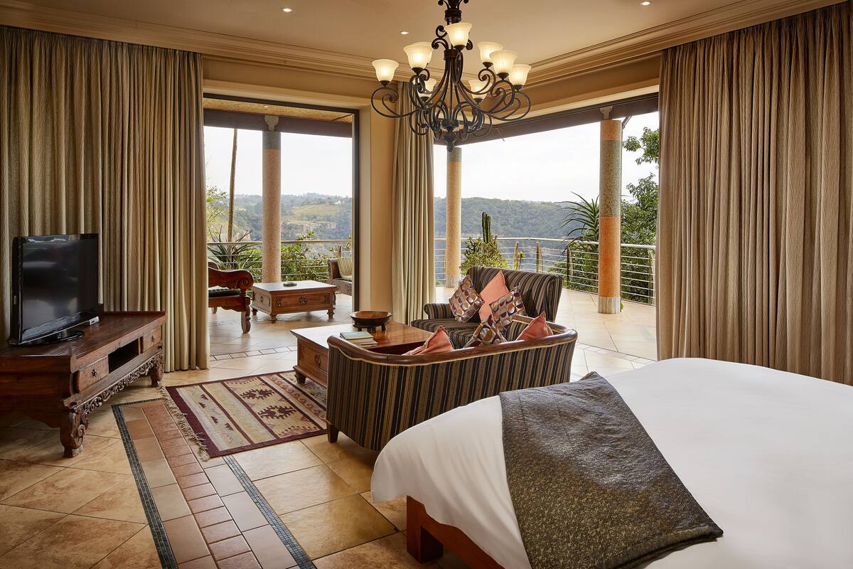 A spacious bedroom with a large bed, a television, a sofa, and a balcony with a view of the hills.