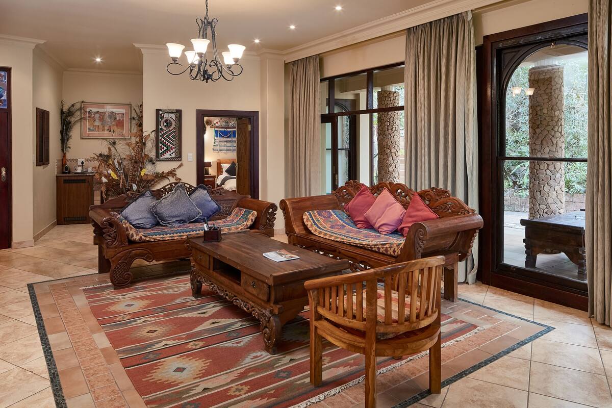 A traditional living room with two sofas, a coffee table, and a wooden chair. There is a large window with curtains, a chandelier, and a stone pillar outside.