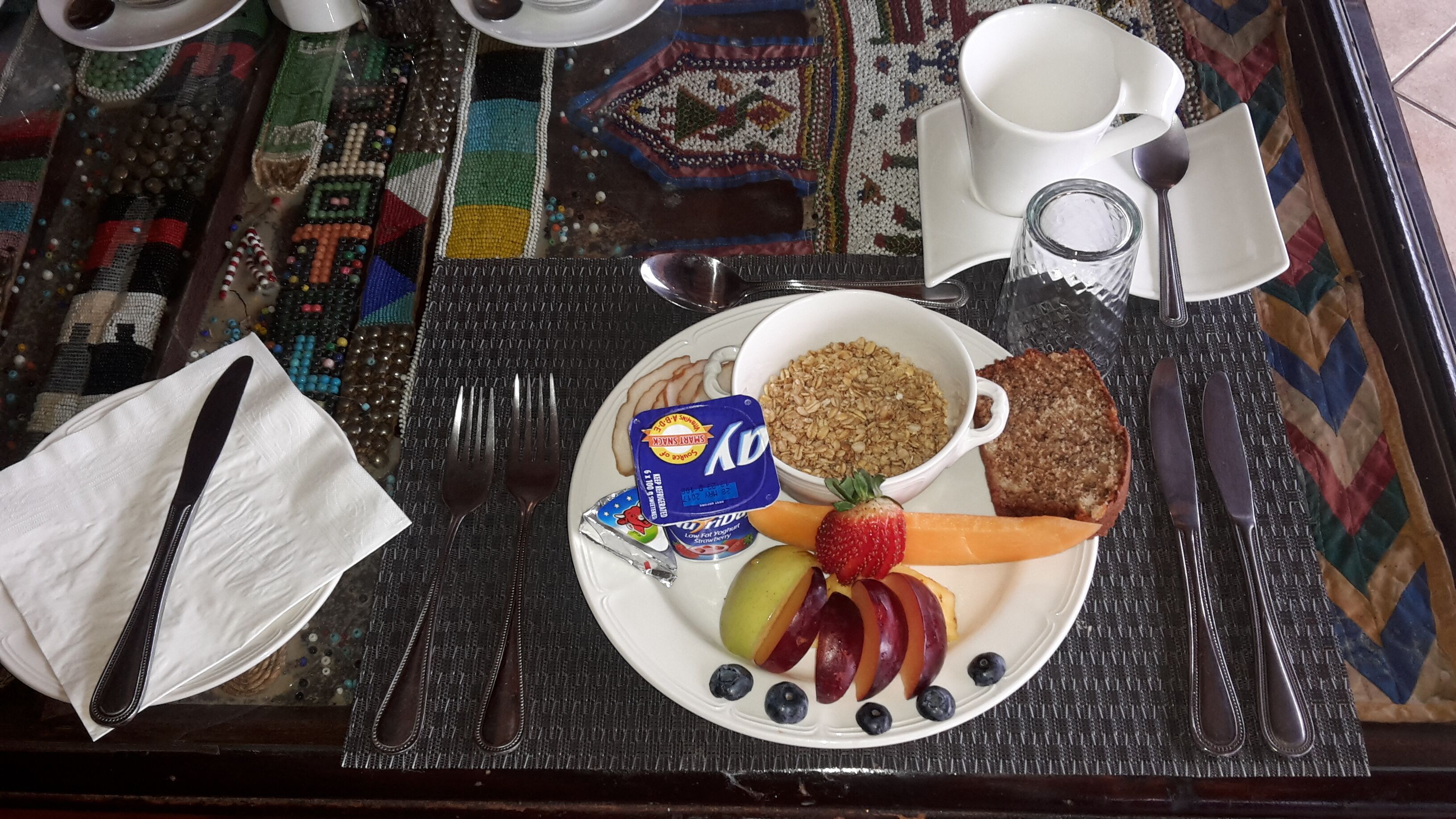 Un desayuno con un plato de yogur, fruta y tostadas, cubiertos y una taza sobre una mesa con un posavasos estampado.