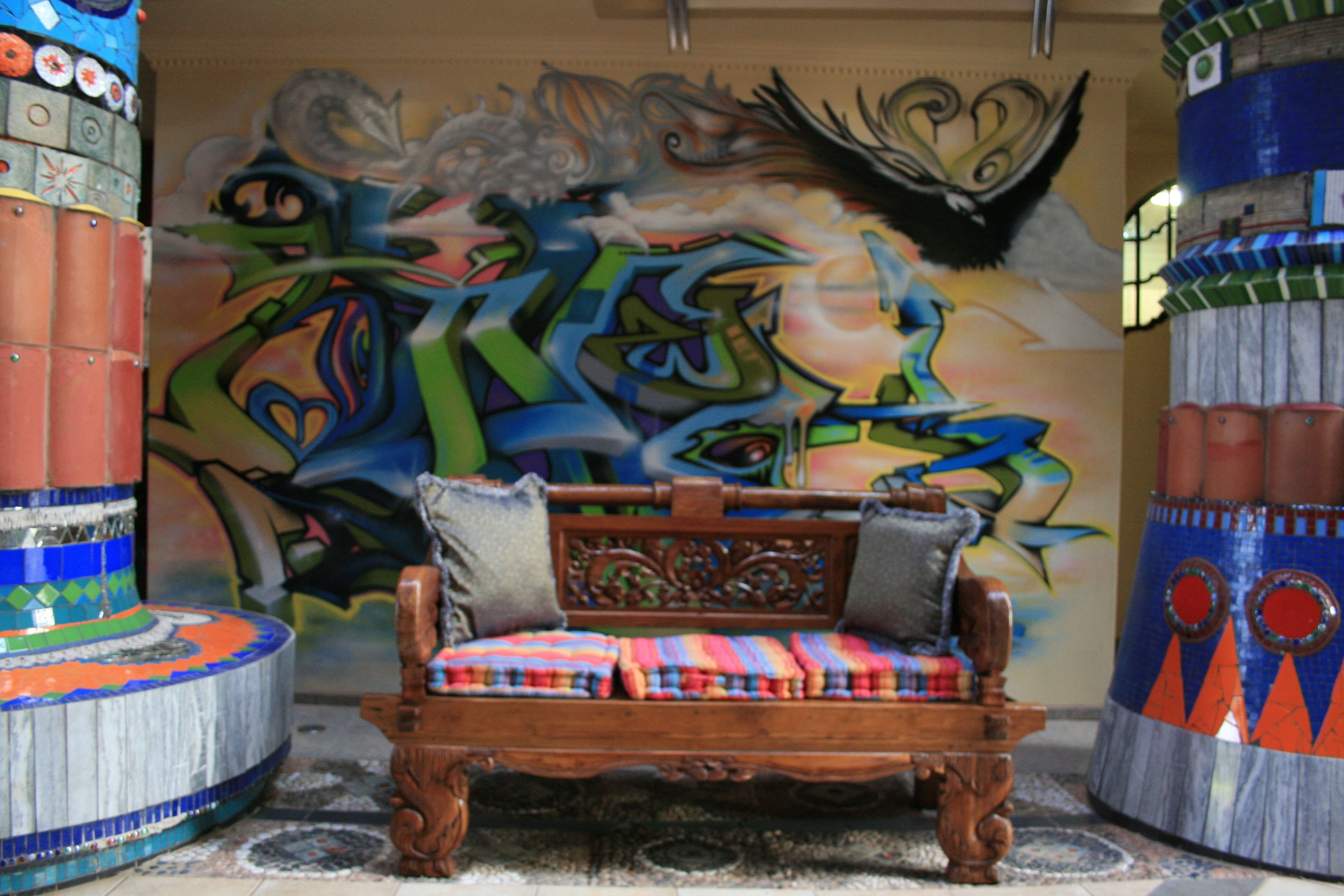 Une pièce avec un banc en bois, des coussins aux couleurs vives et une grande fresque murale abstraite.
