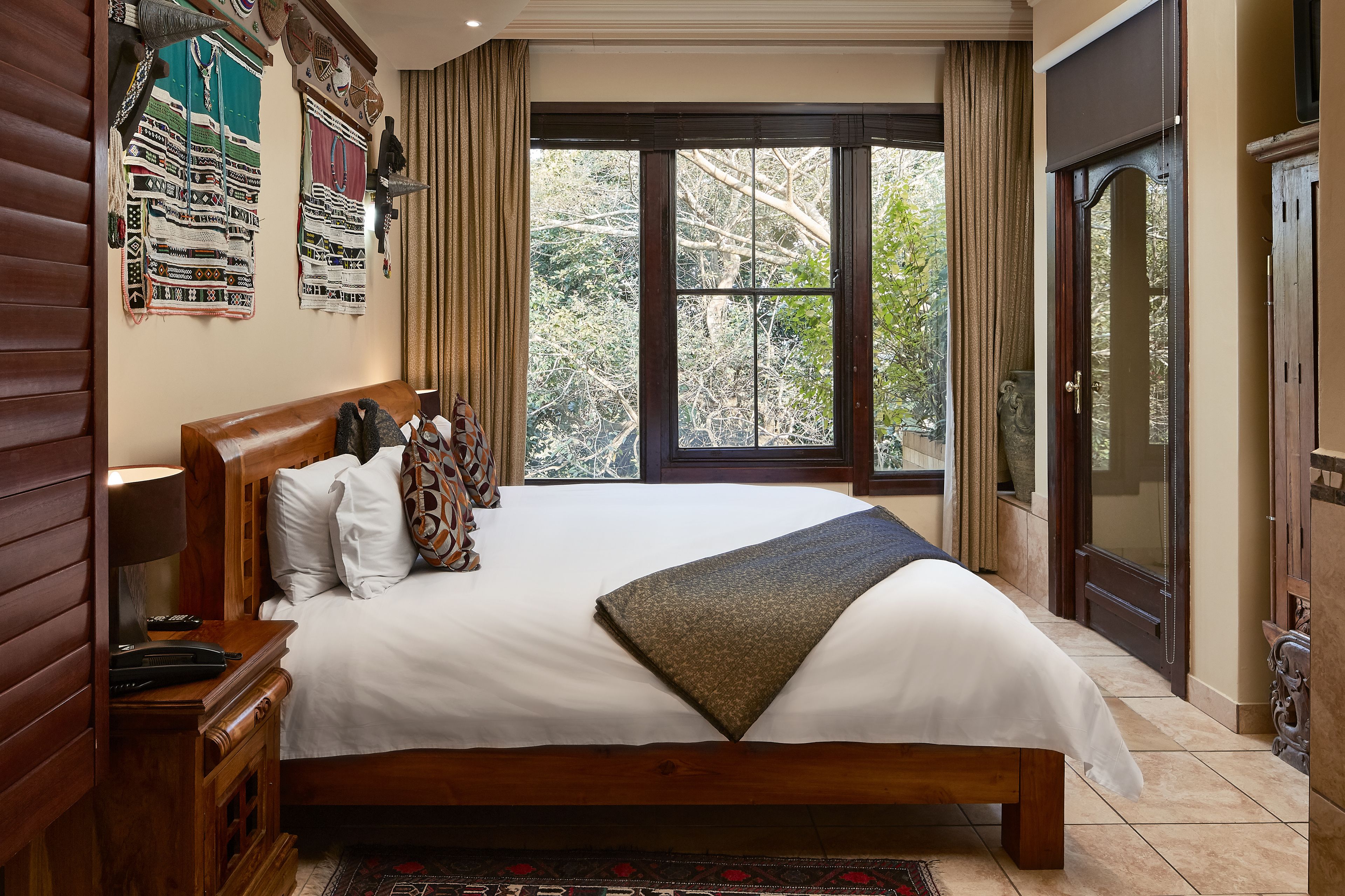 A neatly made bed with a white comforter and a brown throw blanket, wooden headboard, a nightstand with a lamp, and a large window with curtains.
