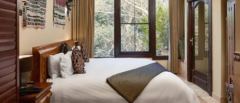 A neatly made bed with a white comforter and a brown throw blanket, wooden headboard, a nightstand with a lamp, and a large window with curtains.