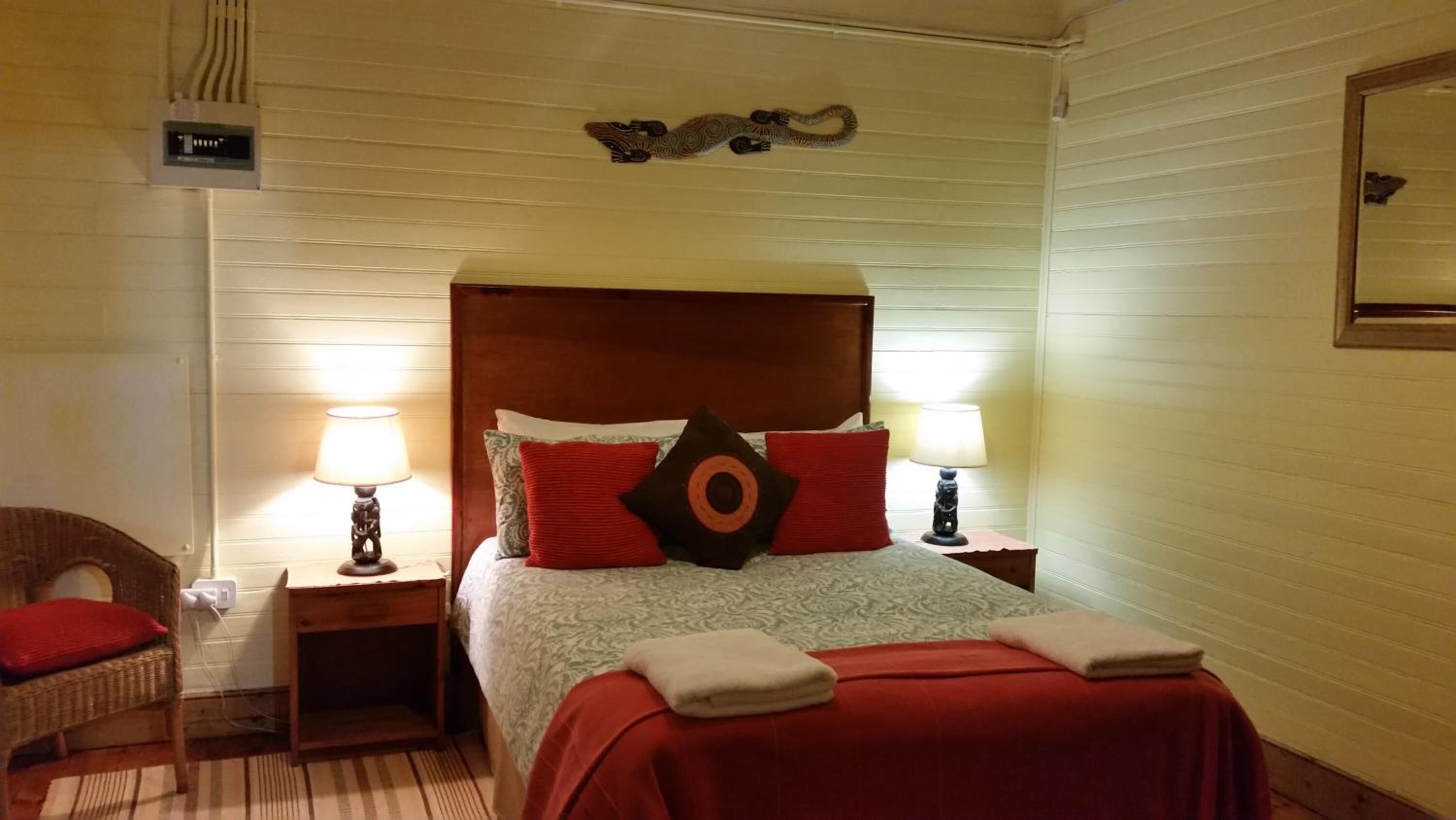 Ein Hotelzimmer mit einem hölzernen Kopfteil, zwei Nachttischlampen, einem Bett mit roten und gemusterten Kissen, einem Korbsessel und einem kleinen Tisch mit einer Lampe.