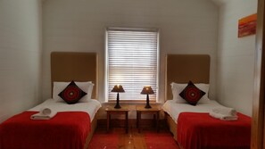 A hotel room with two beds, each with a red bedspread and a decorative pillow, a window with blinds, and two bedside lamps. - Bedrock Lodge (Port Nolloth)