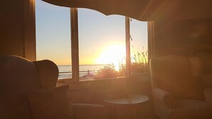 Et værelse med et stort vindue med udsigt over en strand ved solnedgang.