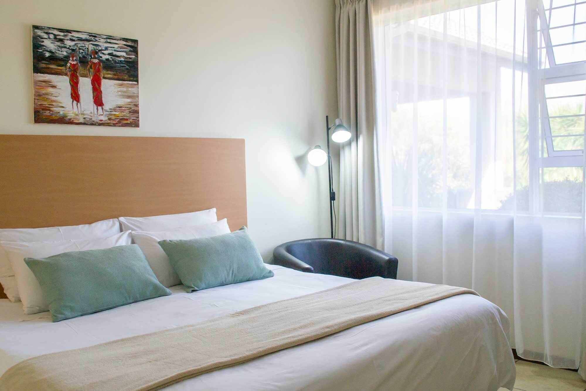 A hotel room with a bed, pillows, a painting on the wall, a chair, and a window with curtains.