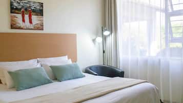 A hotel room with a bed, pillows, a painting on the wall, a chair, and a window with curtains.