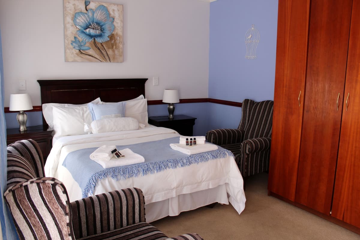 A hotel room with a large bed, two bedside tables with lamps, a striped armchair, a wooden wardrobe, and a floral painting on the wall.