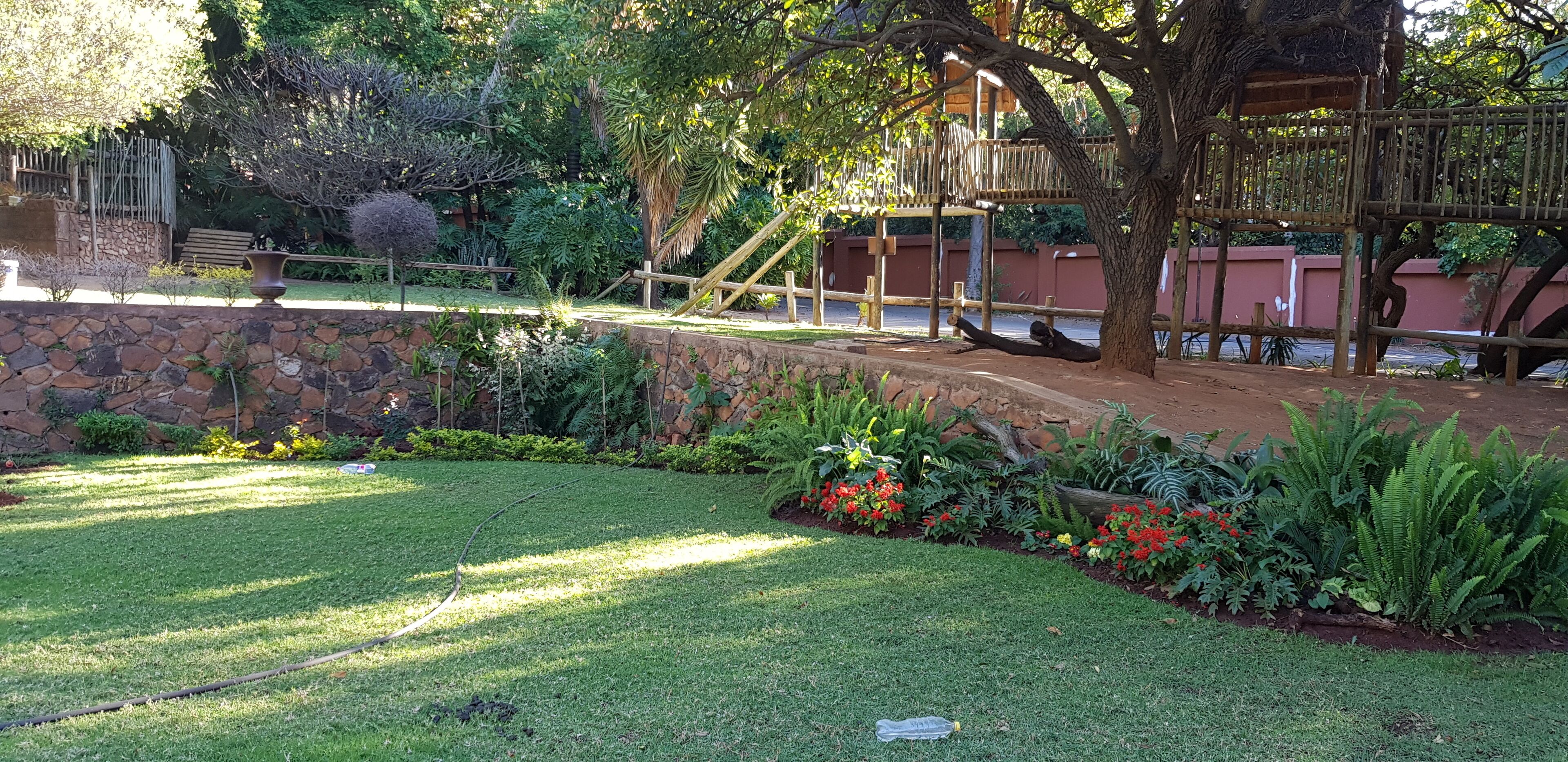Un jardín con muro de piedra, puente de madera y diversas plantas y flores.