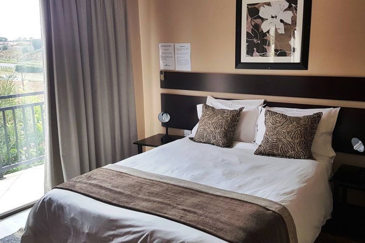 Ein Hotelzimmer mit einem großen Bett, zwei Nachttischen, einem Beistelltisch, einem gerahmten Bild an der Wand und Blick ins Freie durch ein Fenster mit Vorhängen.