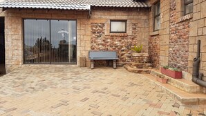 A patio with a brick floor, a bench, a planter with plants, and a building with a tiled roof and stone walls. - Tsessebe Guesthouse (Bloemfontein)