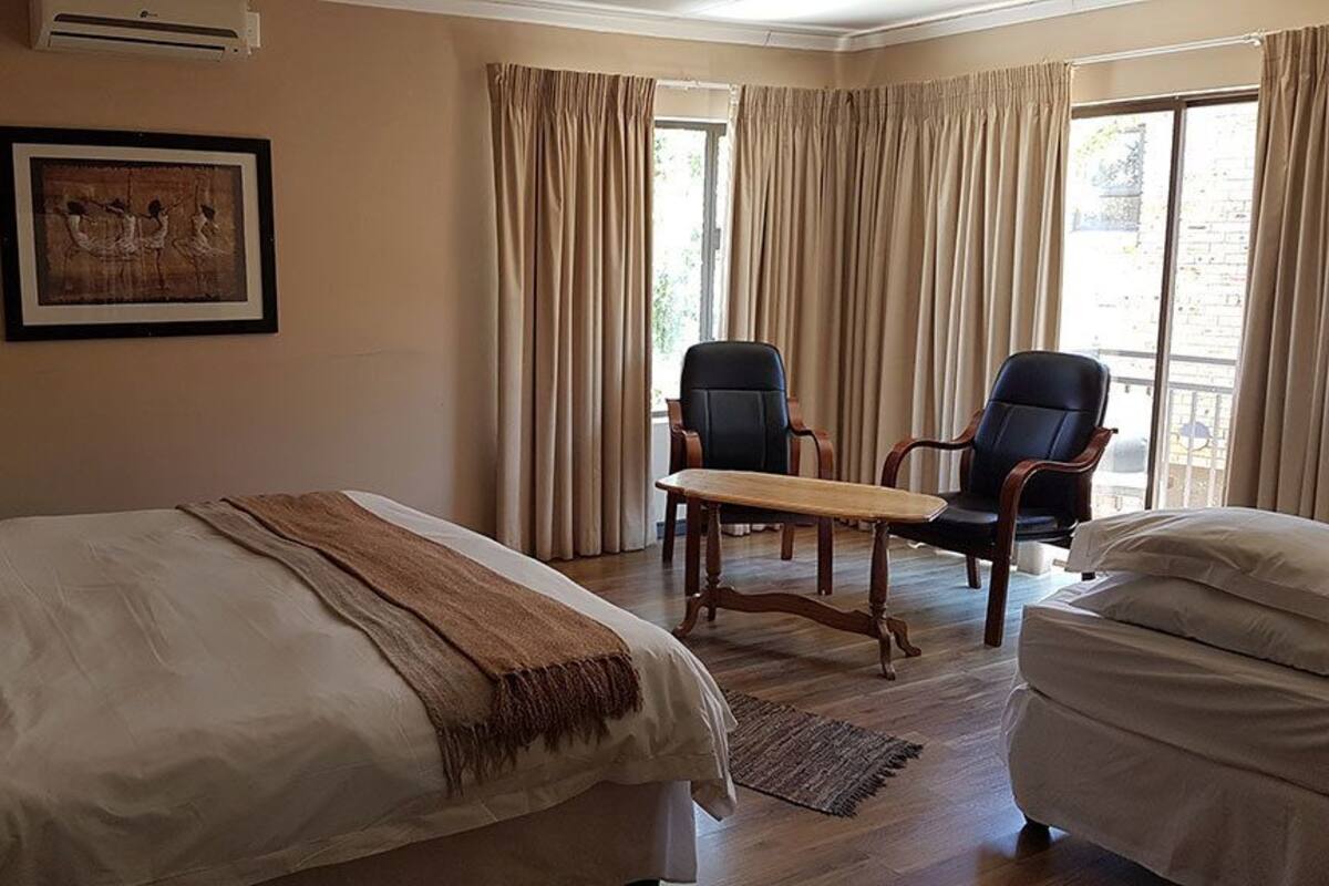 Ein Hotelzimmer mit zwei Betten, einem Holztisch, zwei Stühlen, einem großen Fenster mit Vorhängen und einem gerahmten Bild an der Wand.