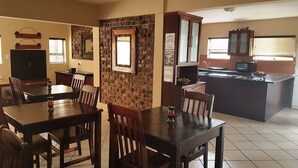 A dining area with a brick wall, wooden tables, and chairs. There is a kitchen area with a refrigerator, microwave, and cabinets. - Tsessebe Guesthouse (Bloemfontein)