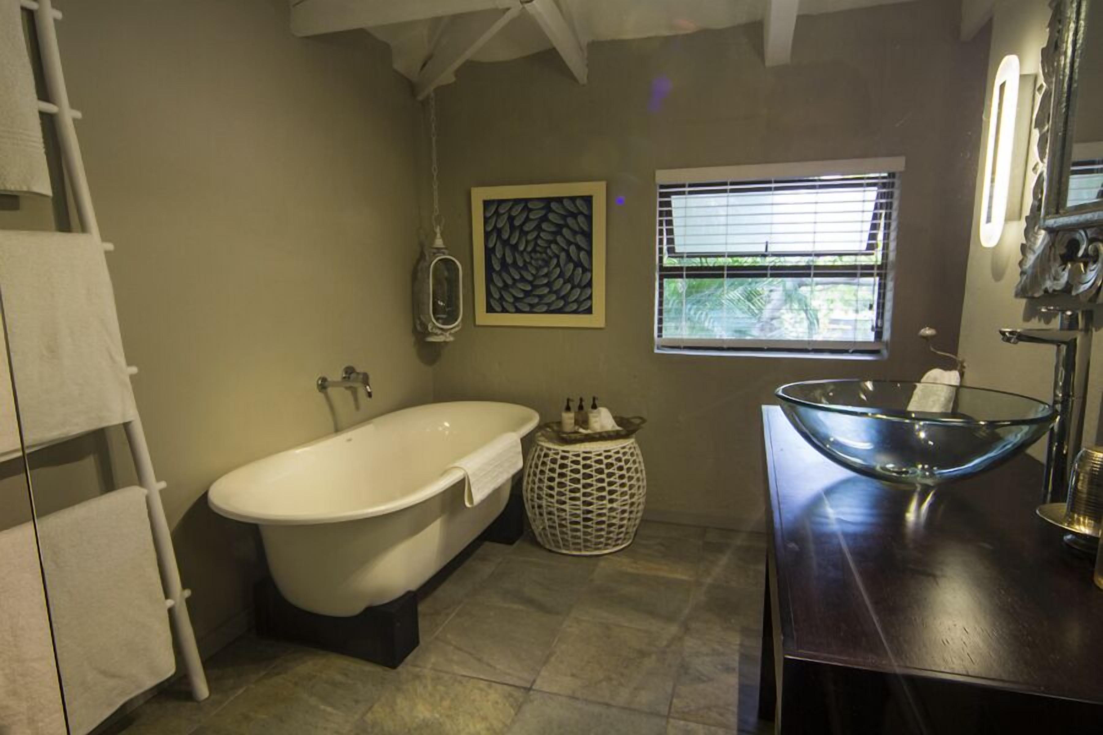 Un baño con una bañera independiente, un lavabo con recipiente de vidrio, un perchero, un adorno de pared y una ventana con persianas.