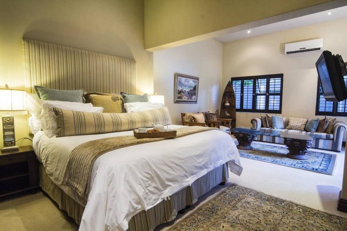 Une chambre spacieuse avec un grand lit, un coin salon avec un canapé et des fauteuils, et une vue sur l’extérieur par les fenêtres.
