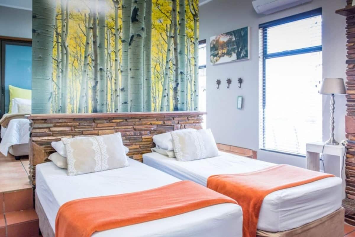 A hotel room with two beds, a stone headboard, a painting of trees, a window with blinds, and a lamp.