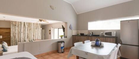 A compact room with a bed, a dining table with chairs, a kitchen area with a refrigerator, microwave, and kettle, and a ceiling fan.