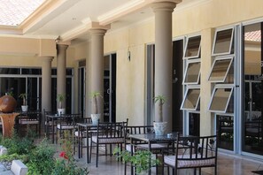 Uma área de pátio externo com várias mesas e cadeiras, colunas grandes e plantas em vasos.