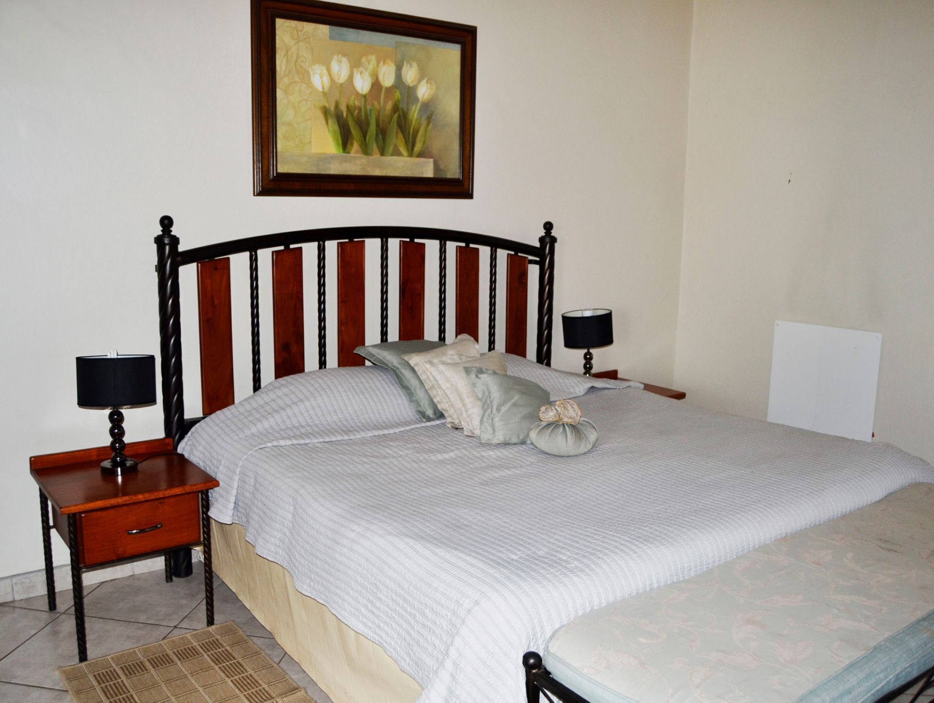 A neatly made bed with a striped bedspread, two bedside tables with lamps, a framed painting above the bed, and a plain wall.