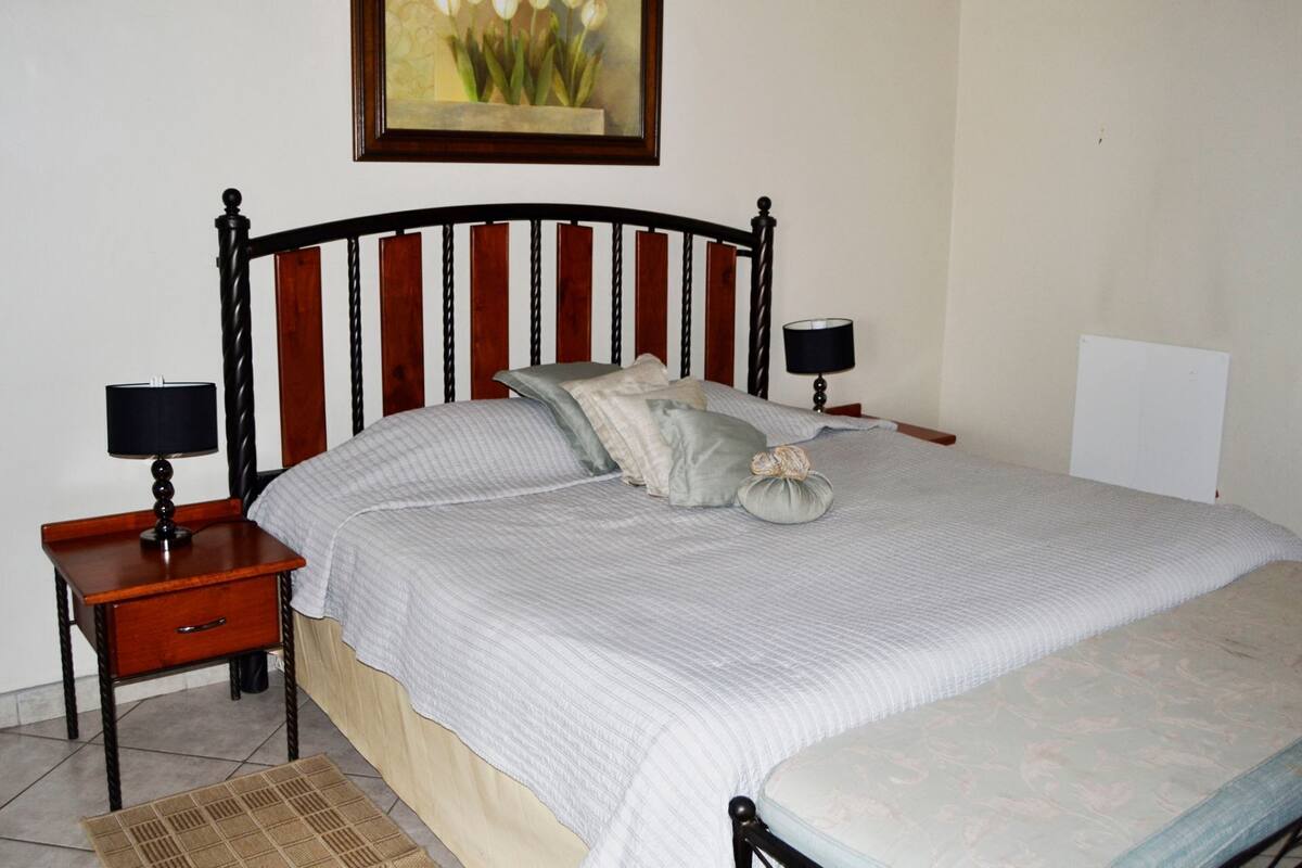 A neatly made bed with a striped bedspread, two bedside tables with lamps, a framed painting above the bed, and a plain wall.
