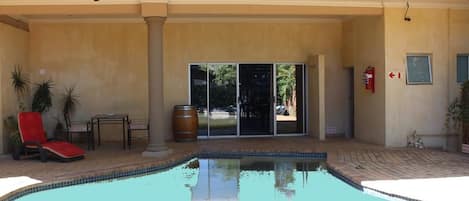 A swimming pool with a red lounge chair and a table, a wooden barrel, and a door leading to a covered area.