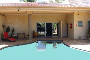 A swimming pool with a red lounge chair and a table, a wooden barrel, and a door leading to a covered area. - Ya Rena Guesthouse (Groblersdal)