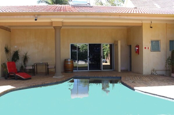 Piscina com uma espreguiçadeira vermelha, uma mesa, um barril de madeira e uma porta que dá para uma área coberta.
