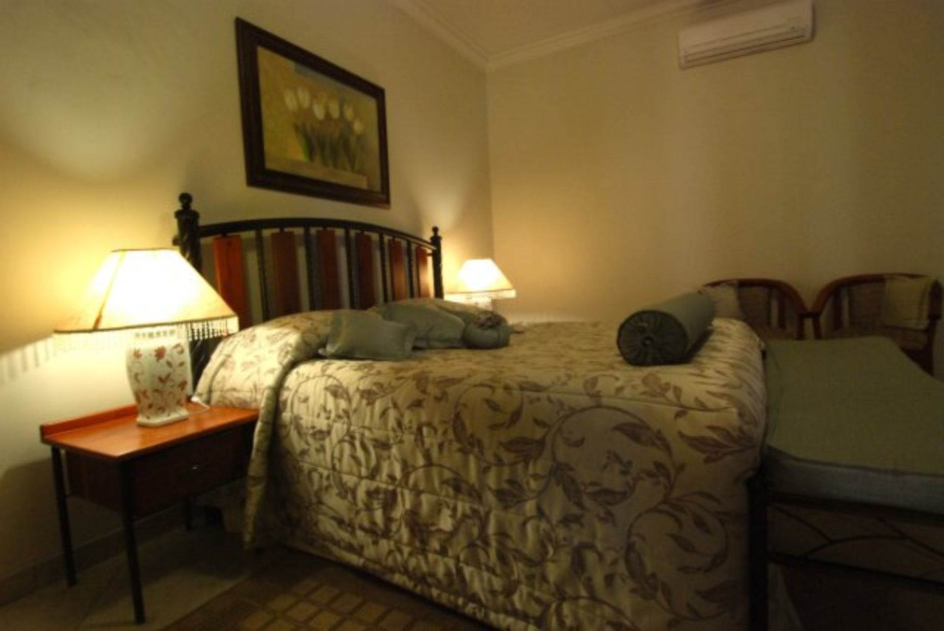 A bedroom with a bed, two bedside lamps, a framed picture on the wall, and an air conditioning unit.