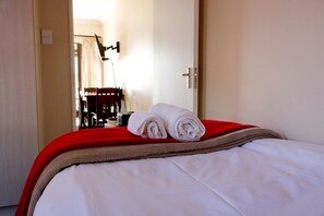 A neatly made bed with white linens and a red blanket, a pair of rolled towels, and a red chair in the background.