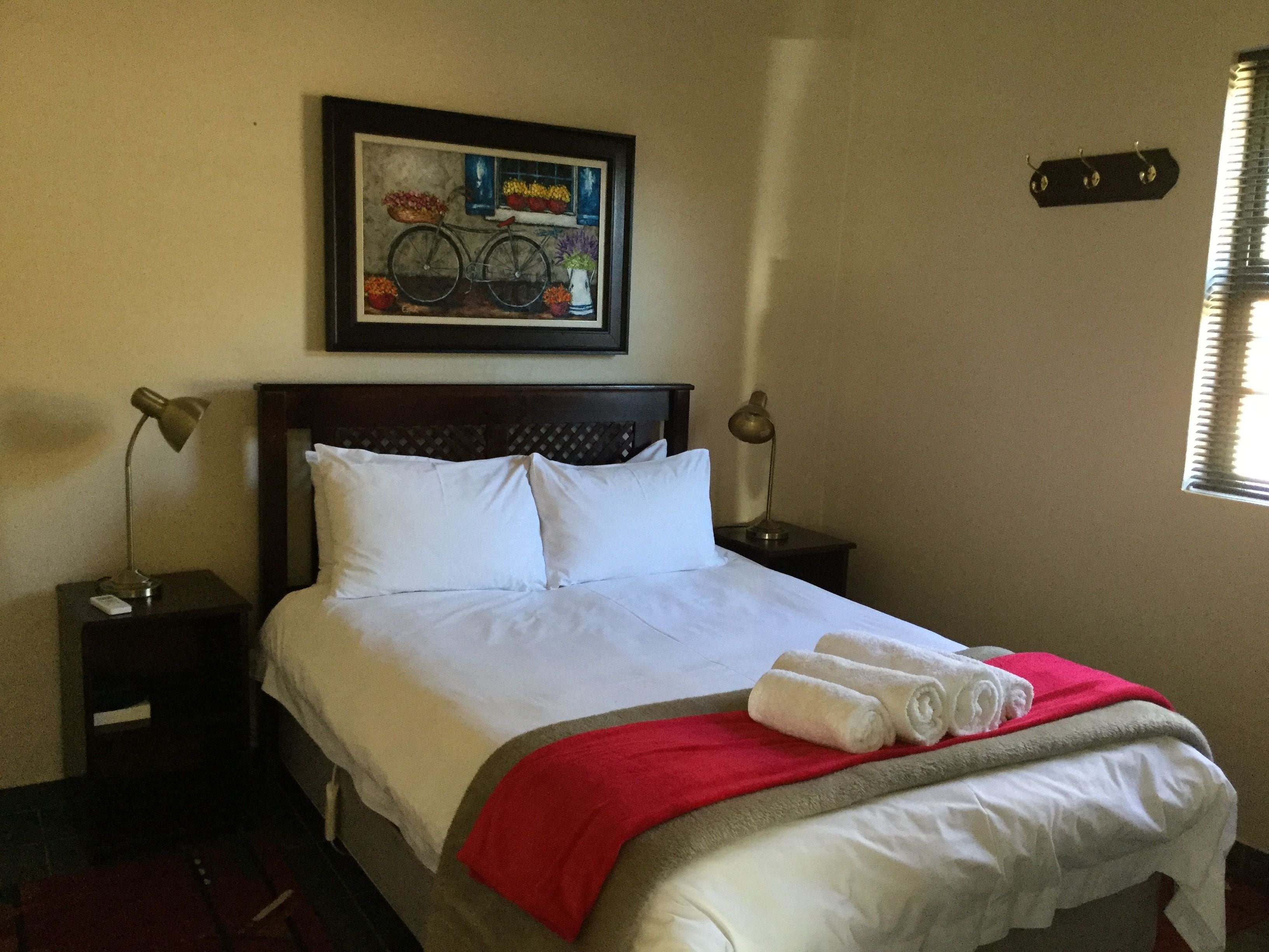 A neatly made bed with white linens and a red blanket, a framed painting above it, and a window with blinds on the right.