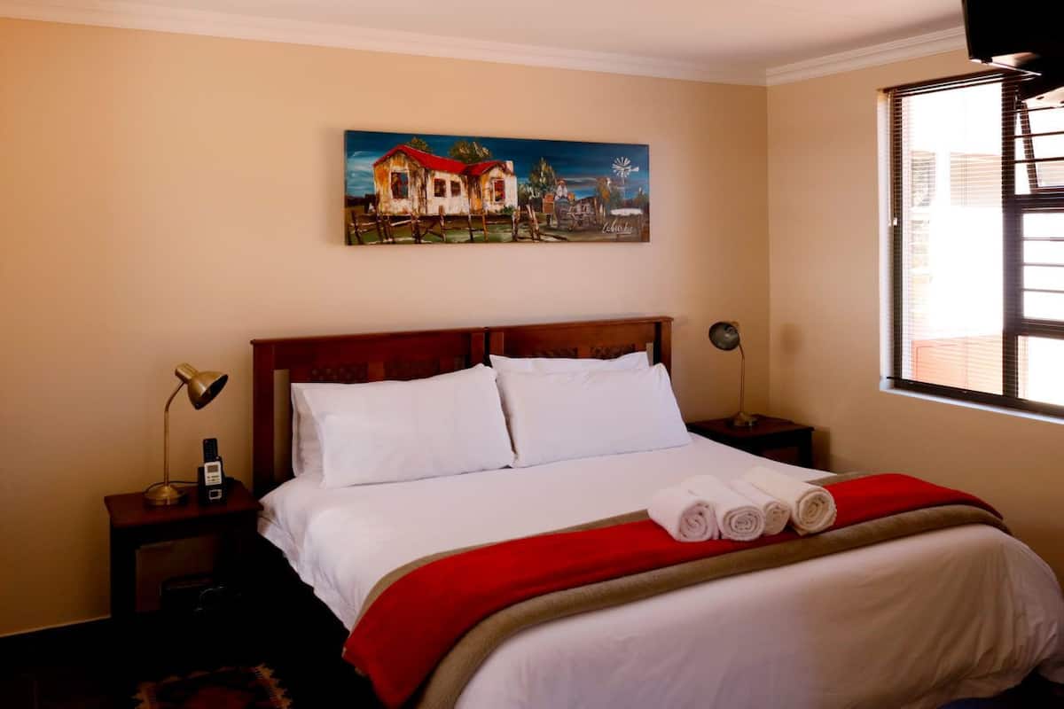 Ein Hotelzimmer mit Bett, Nachttisch mit Lampe, einem Bild an der Wand und einem Fenster mit Jalousien.