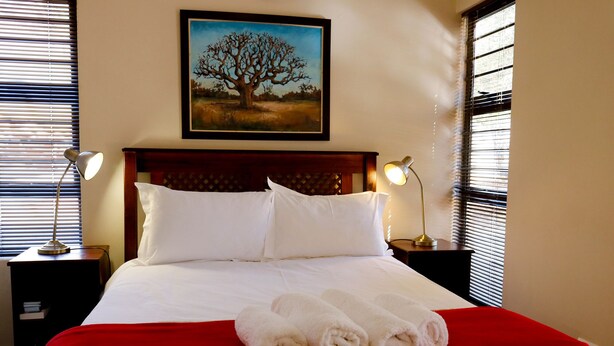 A hotel room with a large bed, two bedside lamps, a framed painting of a tree, and a red bedspread. - A Little Guesthouse (Bloemfontein)