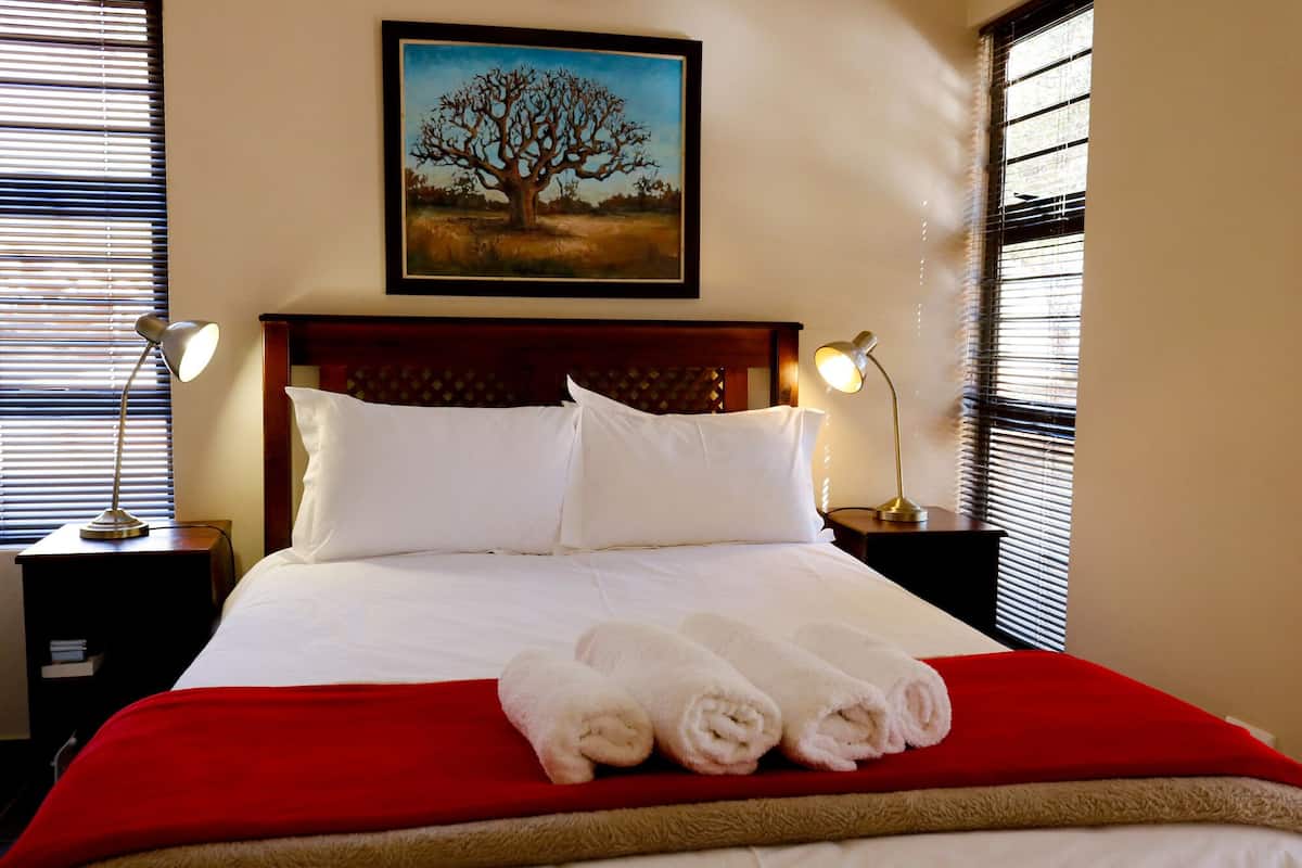 Ein Hotelzimmer mit einem großen Bett, zwei Nachttischlampen, einem gerahmten Bild von einem Baum und einer roten Bettdecke.