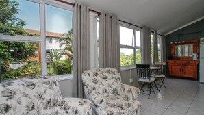 A room with a sofa, a dining table with chairs, a kitchen area, and large windows with curtains. - Beechwood B&B (Durban)